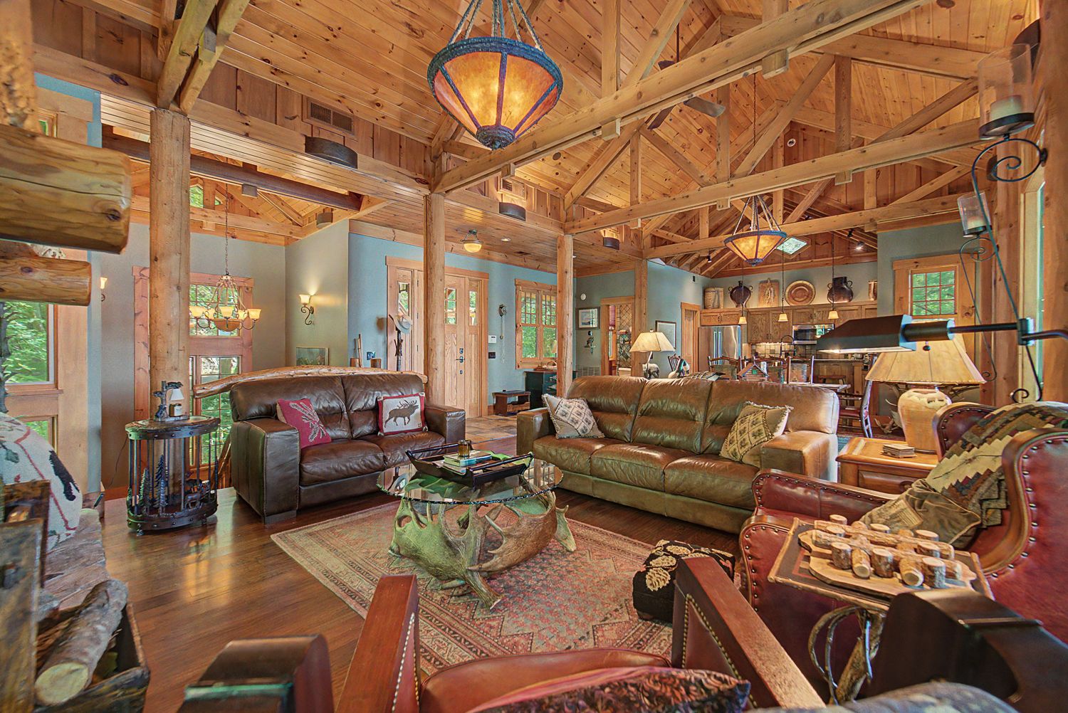 Great room with a vaulted timber-truss ceiling, stacked-stone fireplace, leather seating, and windows on the lake
