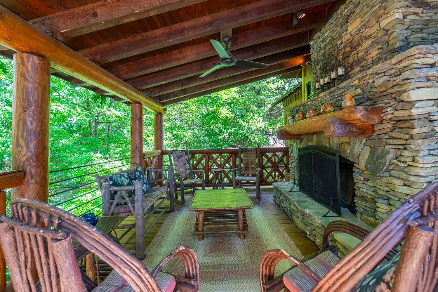 Covered deck with a stacked-stone fireplace, log furniture, and trees on every side