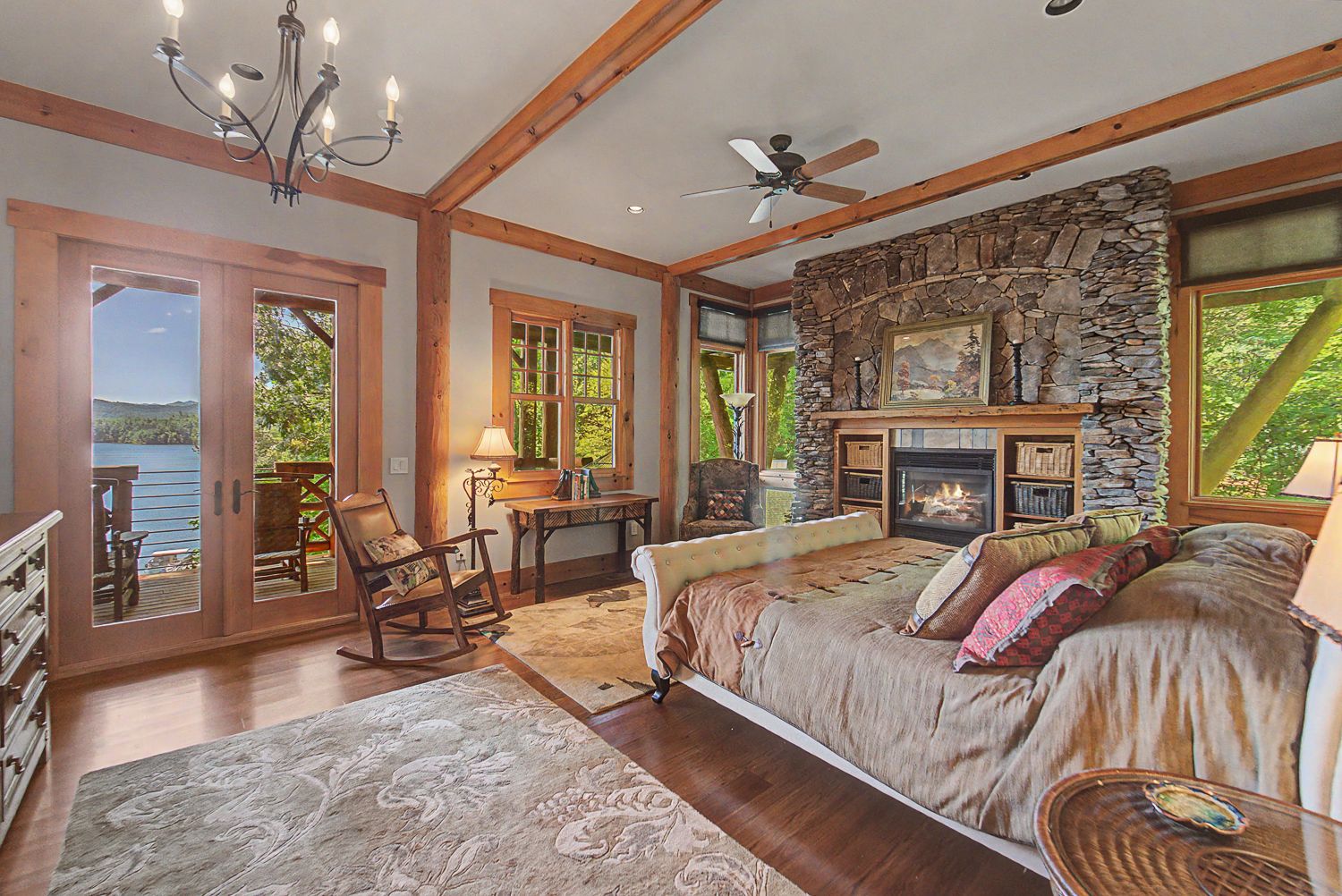 Primary bedroom with a stone fireplace, timber trim, and French doors opening onto a lake balcony